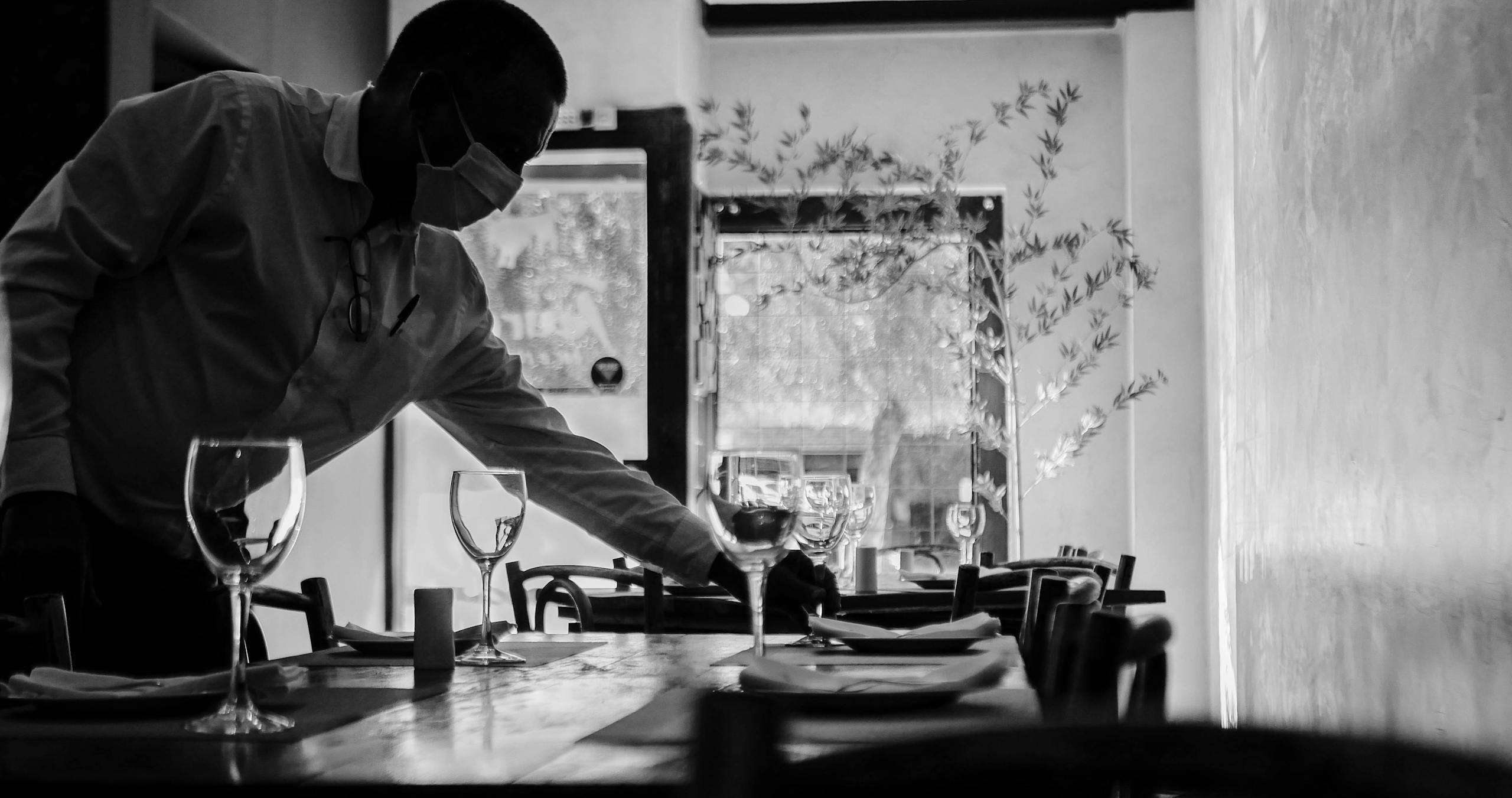 Black and white photo of a masked waiter setting a table in a restaurant.